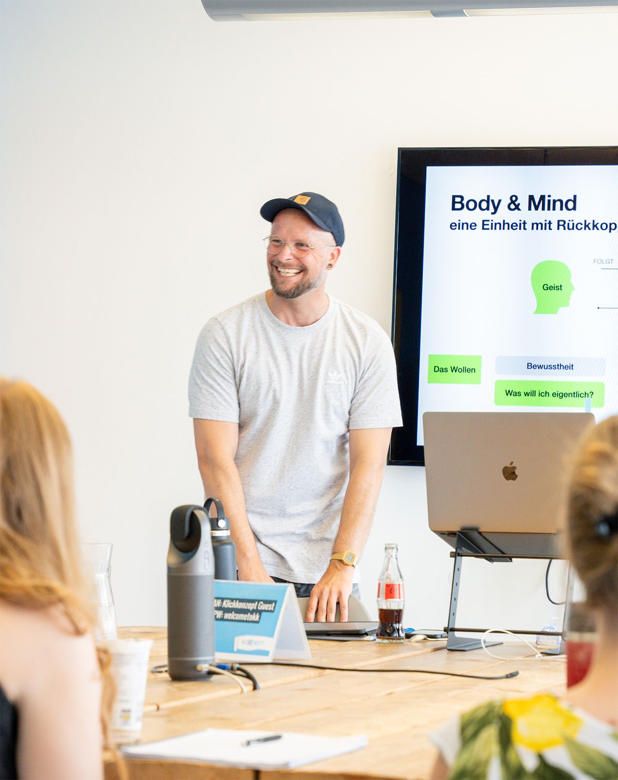 Lächelnder Präsentator mit Kappe leitet Workshop über Body & Mind vor Zuhörern im hellen Seminarraum.