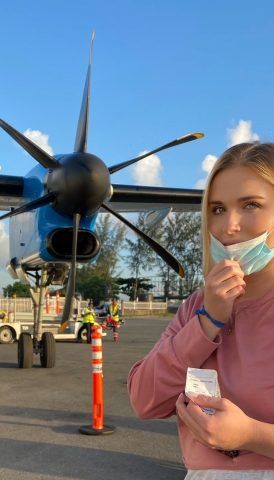 Junge Frau mit abgenommener Gesichtsmaske vor Propellermaschine auf Flugfeld.